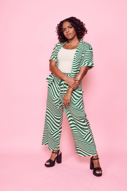 Full-length portrait of a Black woman in a patterned green pantsuit and platform heels, posing in front of a pink background.