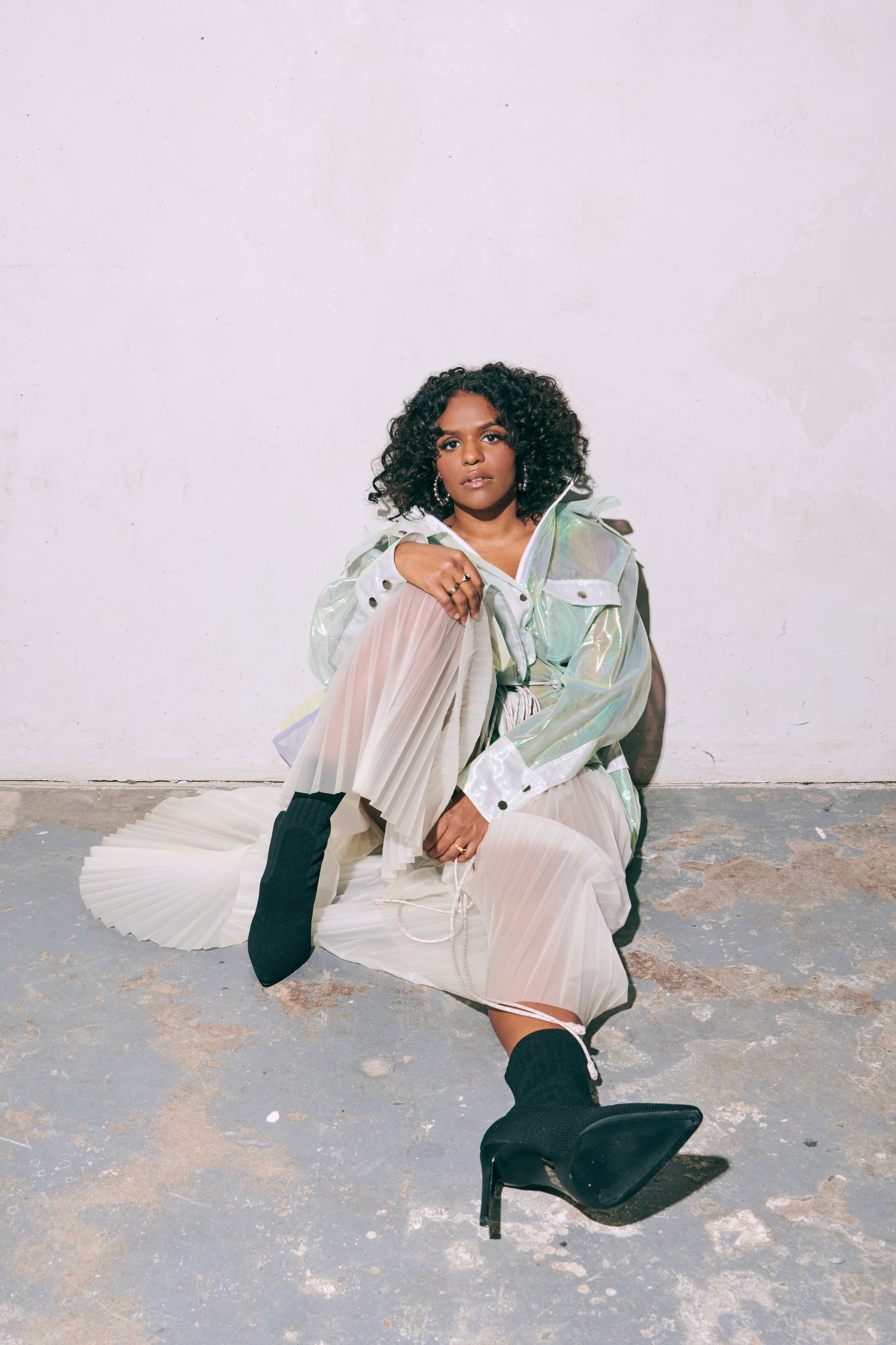 A woman with curly hair sits on the floor in a sheer skirt, iridescent jacket, and black heeled boots against a white background.