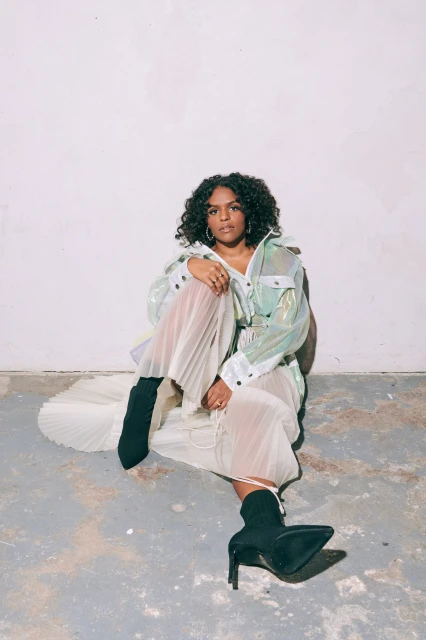 A woman with curly hair sits on the floor in a sheer skirt, iridescent jacket, and black heeled boots against a white background.