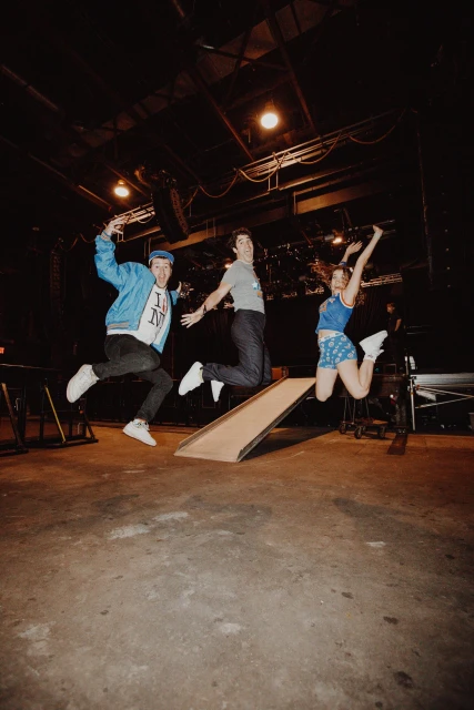 Three young adults energetically jump from a ramp on a stage with visible audio equipment under dark, diffused lighting conditions indoors.