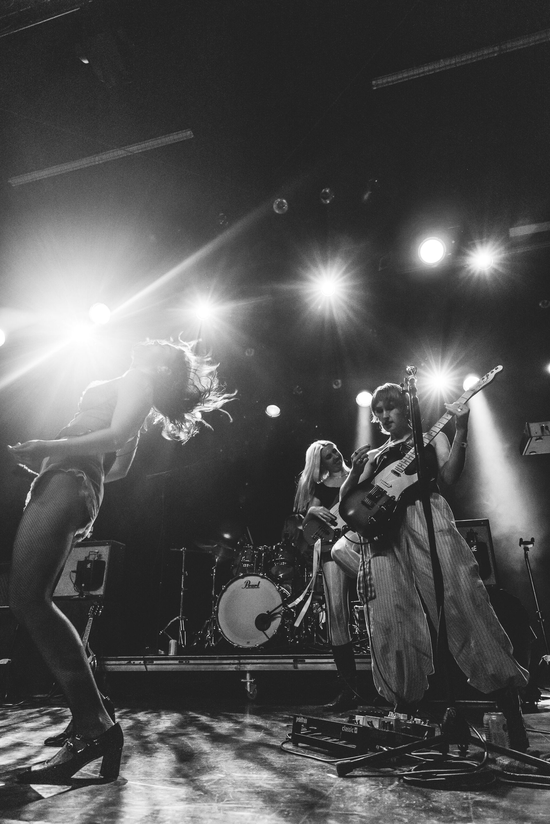 Three musicians perform on a concert stage with strong backlighting, drums, and floor lighting in a high-energy monochrome performance.