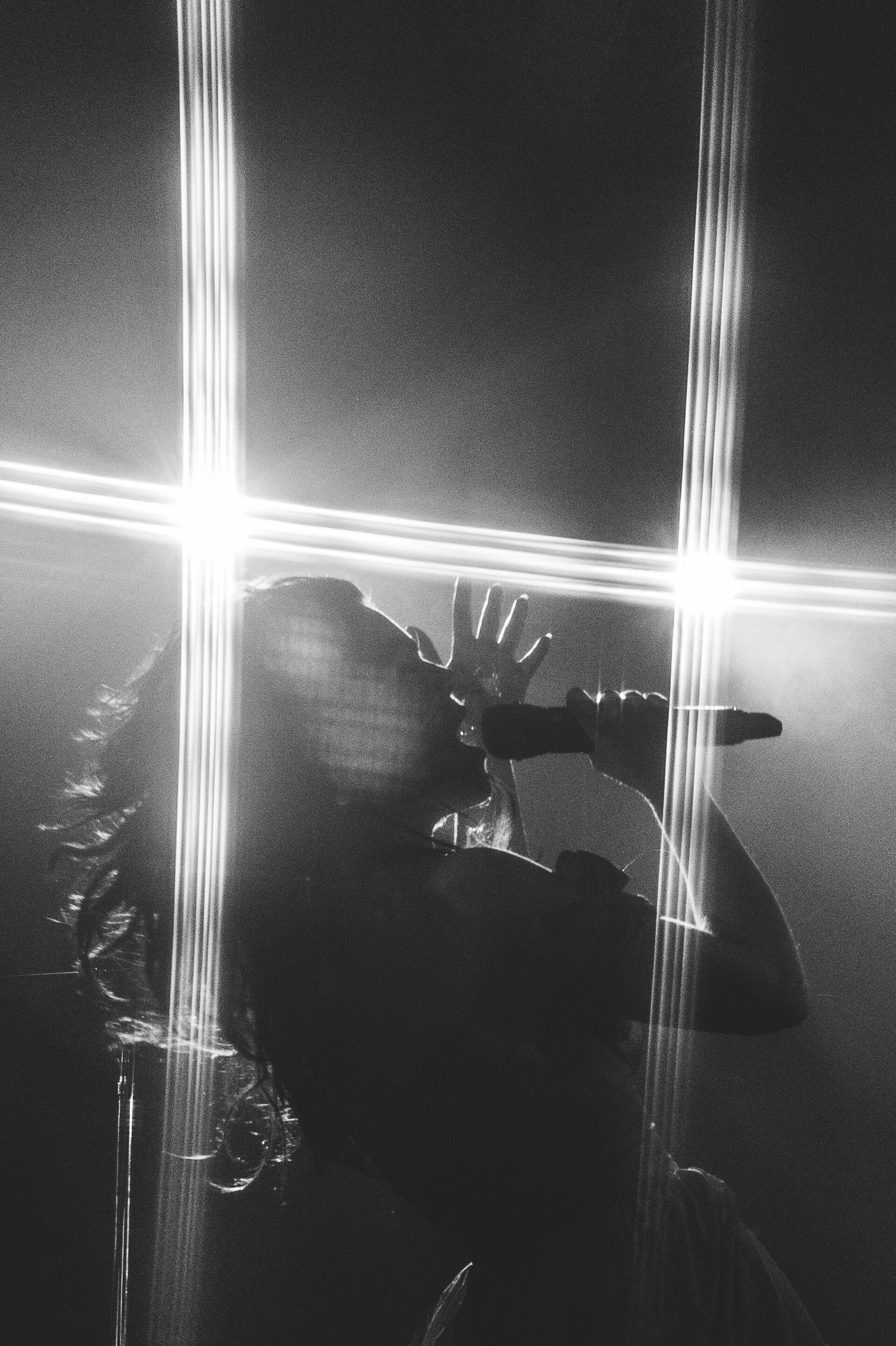Monochrome shot of a vocalist performing on stage, holding a microphone and illuminated by bright lights, captured in a vertical composition.