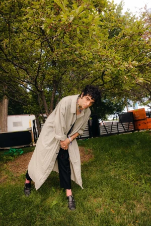 Adult male in a trench coat stands in a grassy park under a tree. He wears dark pants and shoes, a silver chain, and a bracelet.