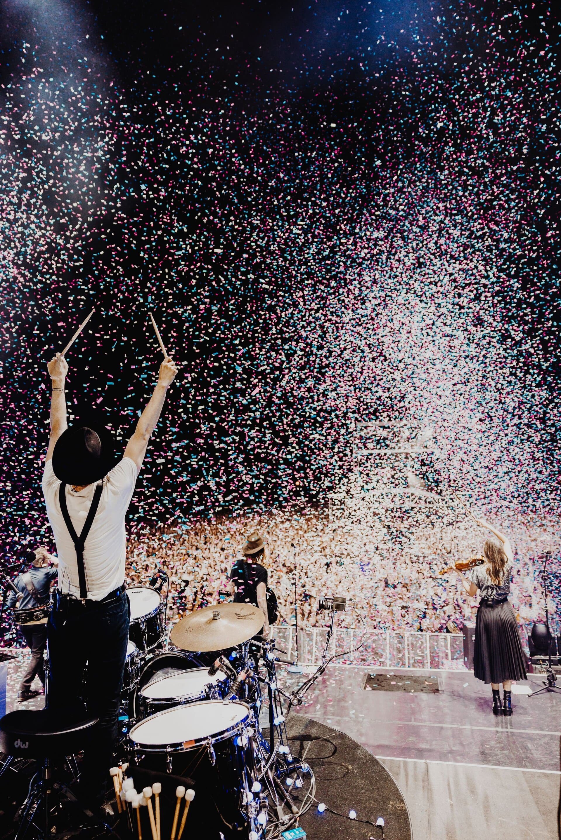 Concert stage with drummer raising sticks amid confetti, violin player, and band members performing for a large crowd in a bright venue.