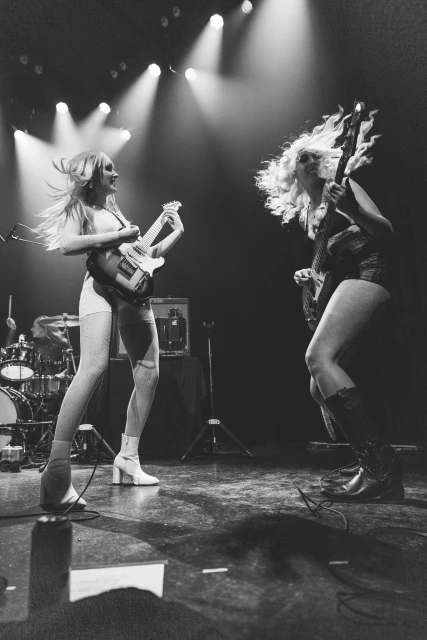 Two women perform on stage with guitars under bright lights, showcasing a live music event in a monochromatic stage setting.