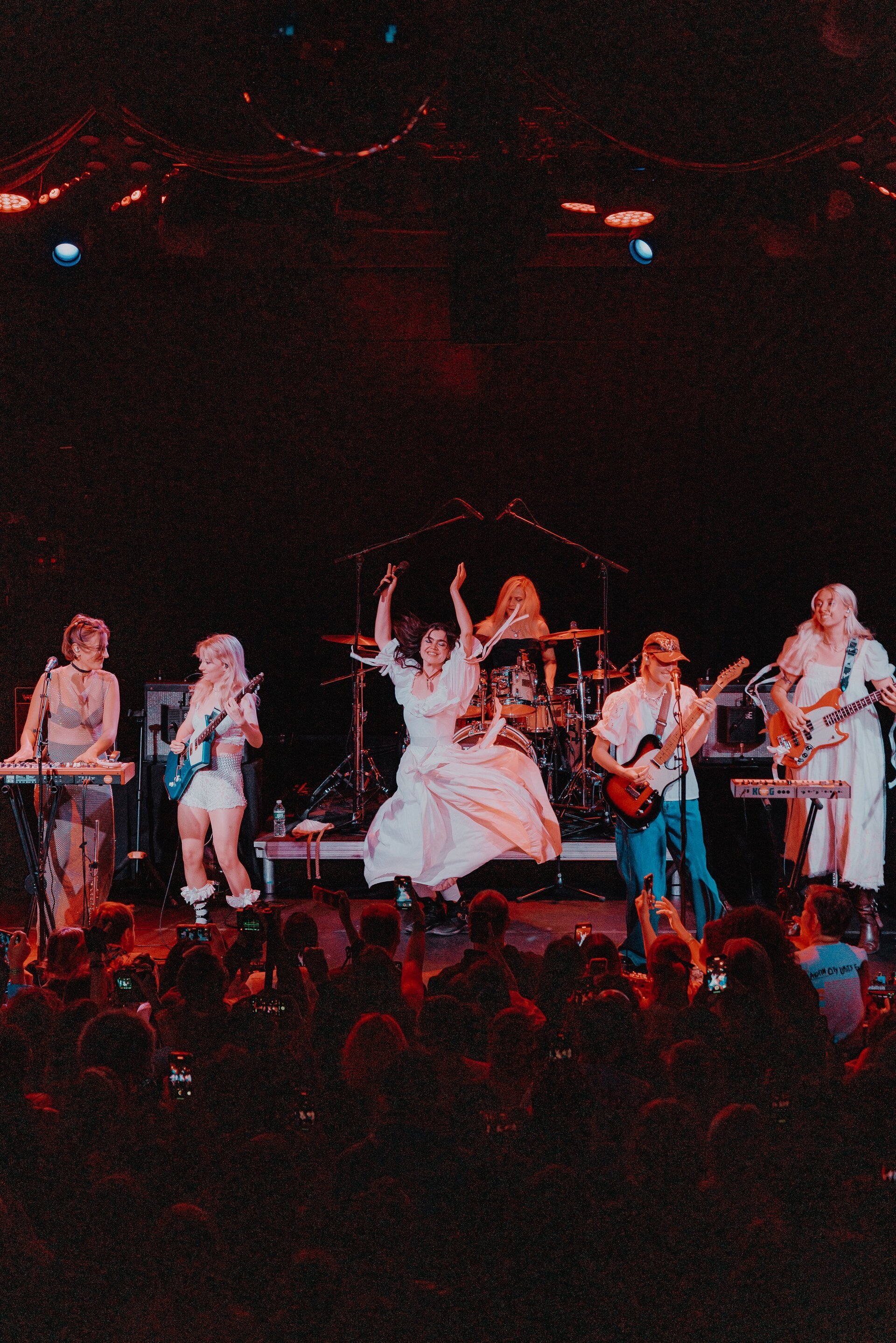 Band performing on stage in concert to a crowd with red stage lighting, drummer, singer, keyboard, and guitar players.