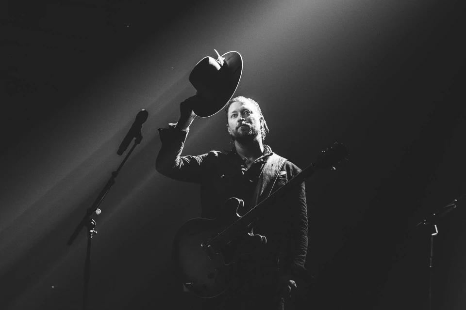 Monochrome shot of a musician on stage holding a hat, wearing a jacket, with an electric guitar and microphone stand in view.