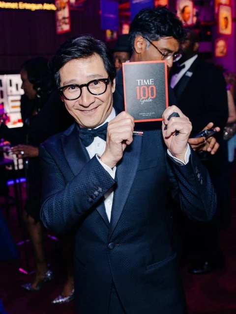 Smiling man in tuxedo holds Time 100 Gala program at indoor event with blurred figures in background. He wears glasses and a bowtie.