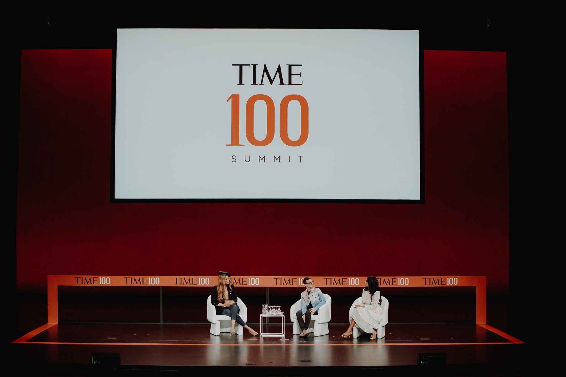 Panel discussion featuring three seated individuals on a stage with a screen displaying the TIME 100 Summit logo in a well-lit indoor venue.