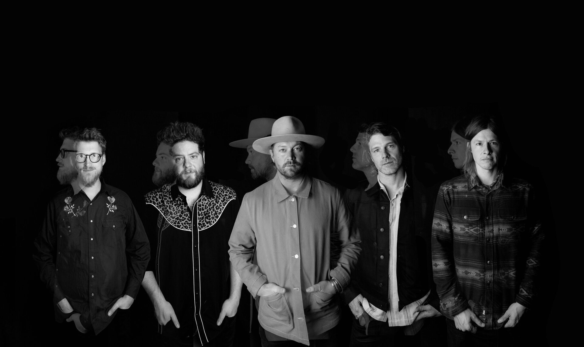 Black and white studio shot of five adult males, appearing as a band, wearing casual attire and standing in front of a dark background.