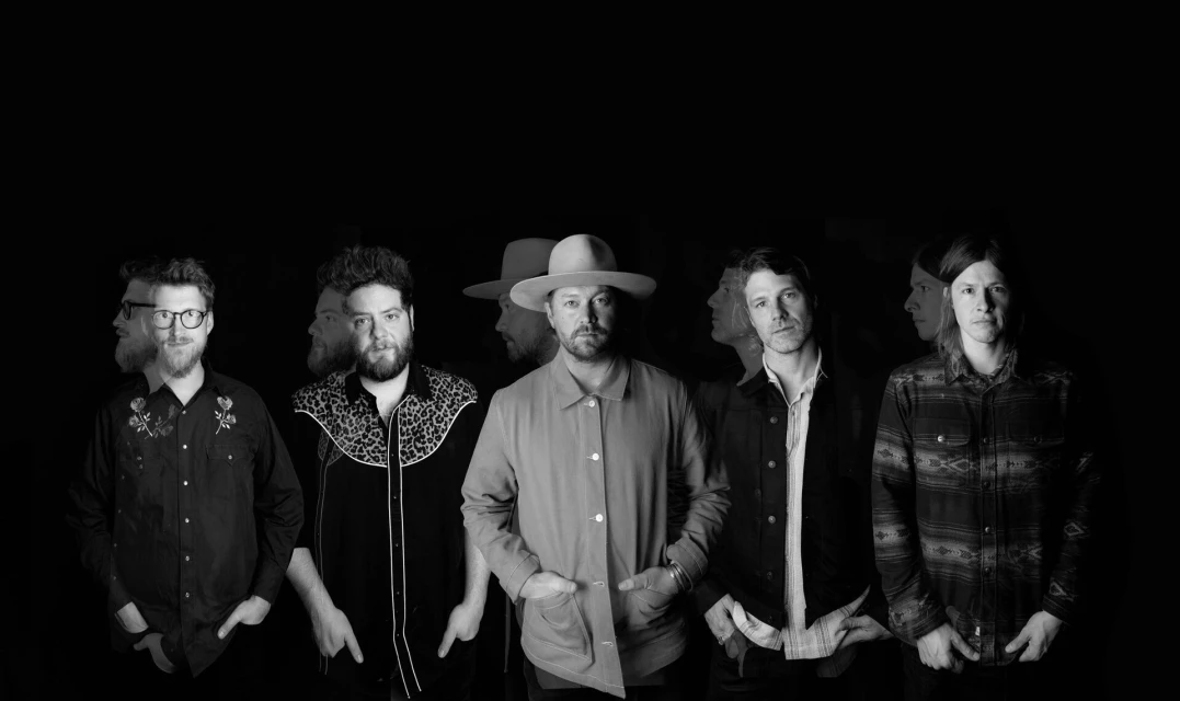 Black and white studio shot of five adult males, appearing as a band, wearing casual attire and standing in front of a dark background.