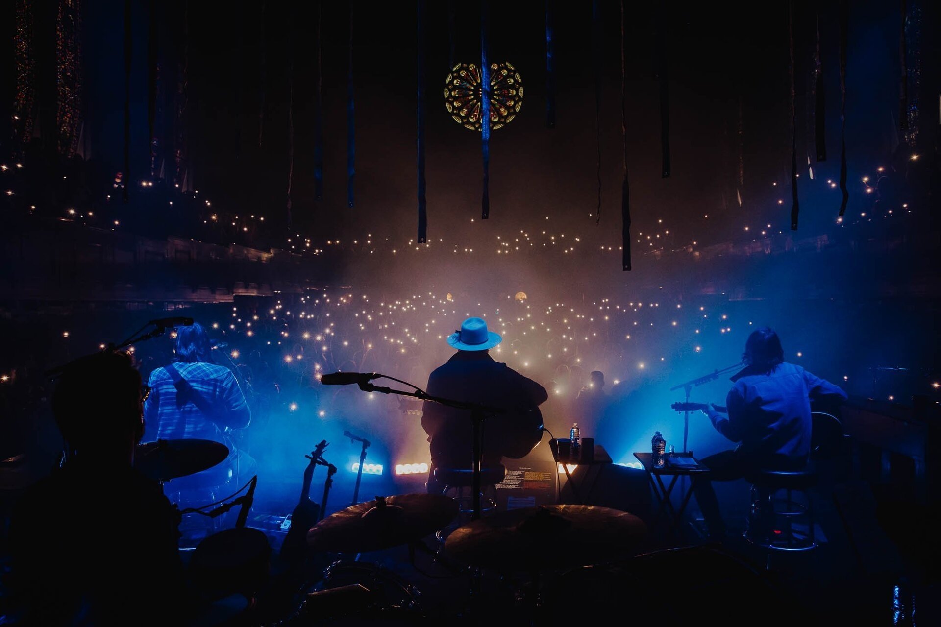 A live music performance features musicians playing instruments on stage in a dark venue with illuminated spotlights and a cheering audience.