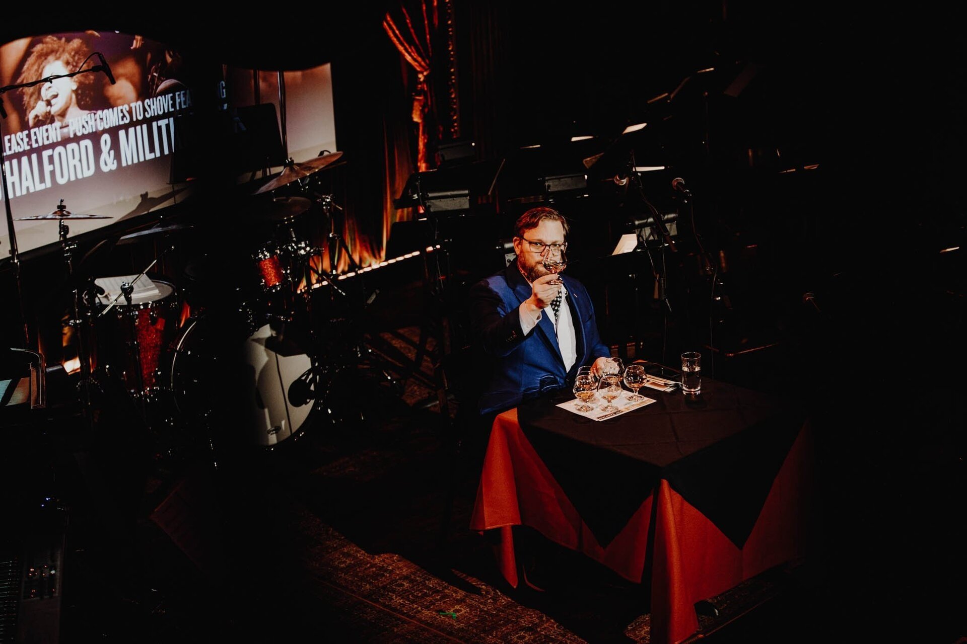 Adult male in a blue blazer sitting at a table with glasses in a dimly lit concert venue with band instruments and stage lighting.