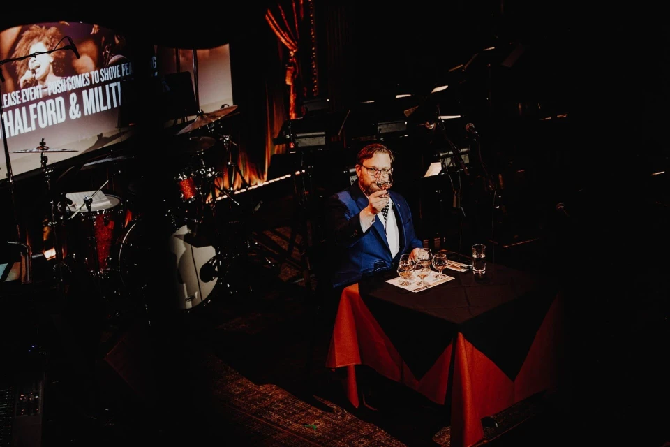 Adult male in a blue blazer sitting at a table with glasses in a dimly lit concert venue with band instruments and stage lighting.