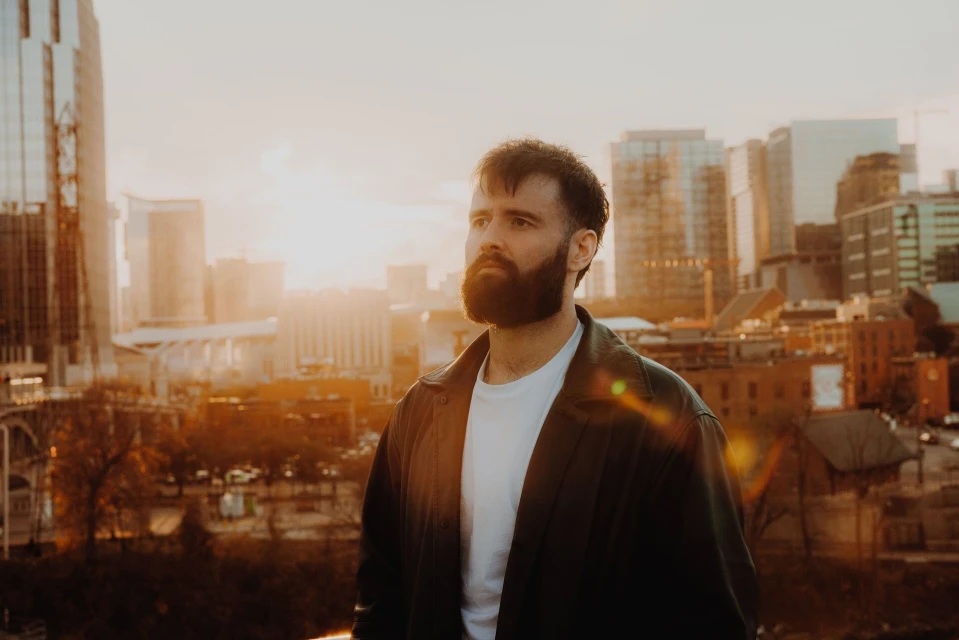 Man with a beard wearing a jacket and white t-shirt standing on a rooftop in an urban environment with city buildings in the background.