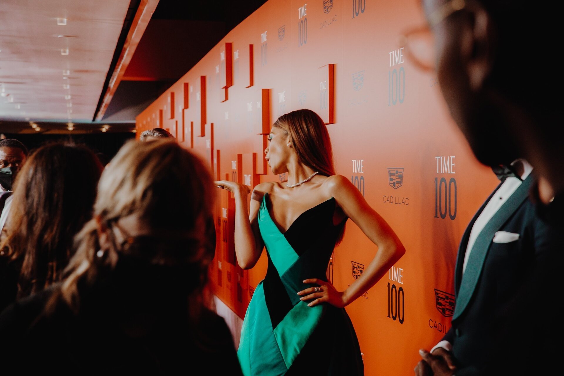 A woman in a green and black dress stands on a red carpet with the TIME 100 logo, posing for a photo while blowing a kiss to the camera.