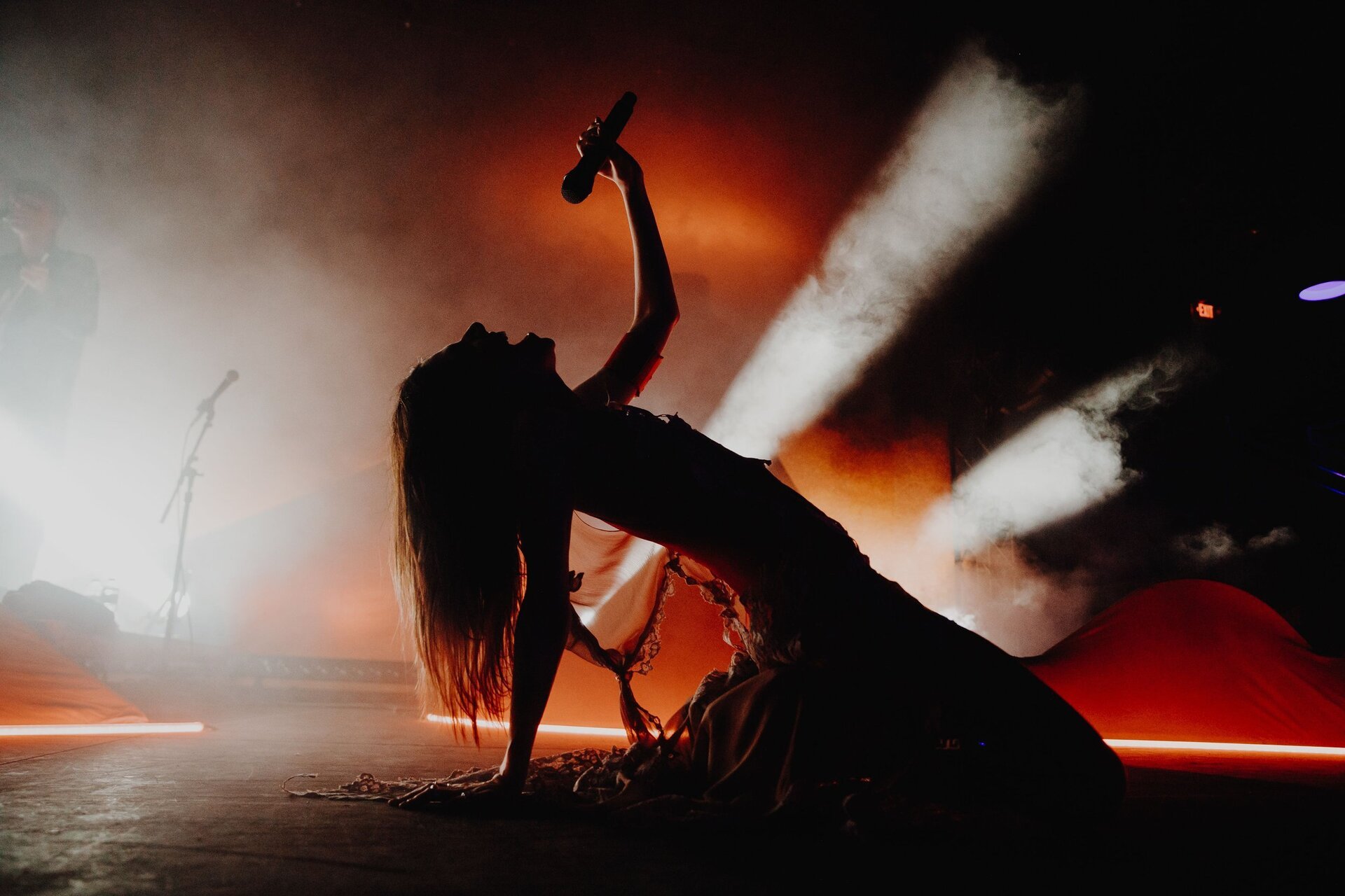 Silhouette of a female singer on stage with a microphone, lit by bright spotlights and orange stage lighting during a concert.