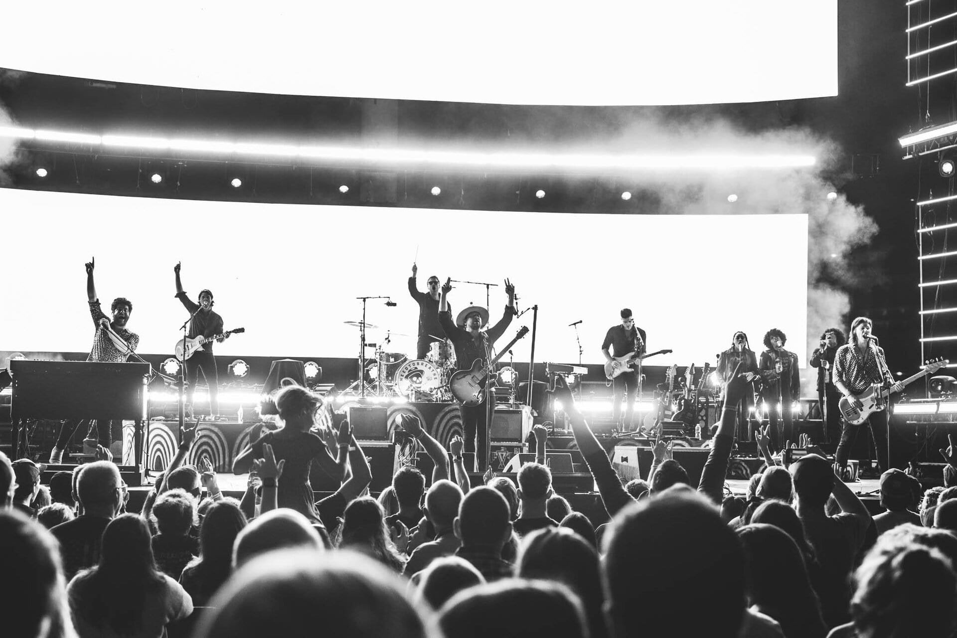 Black and white shot of a live concert featuring a band performing on stage before a large crowd in a brightly lit venue.