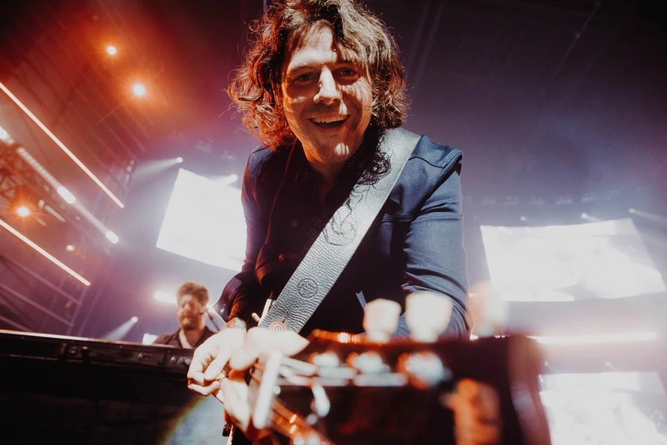 Adult male musician playing guitar on stage with a keyboardist visible in the background and stage lighting.