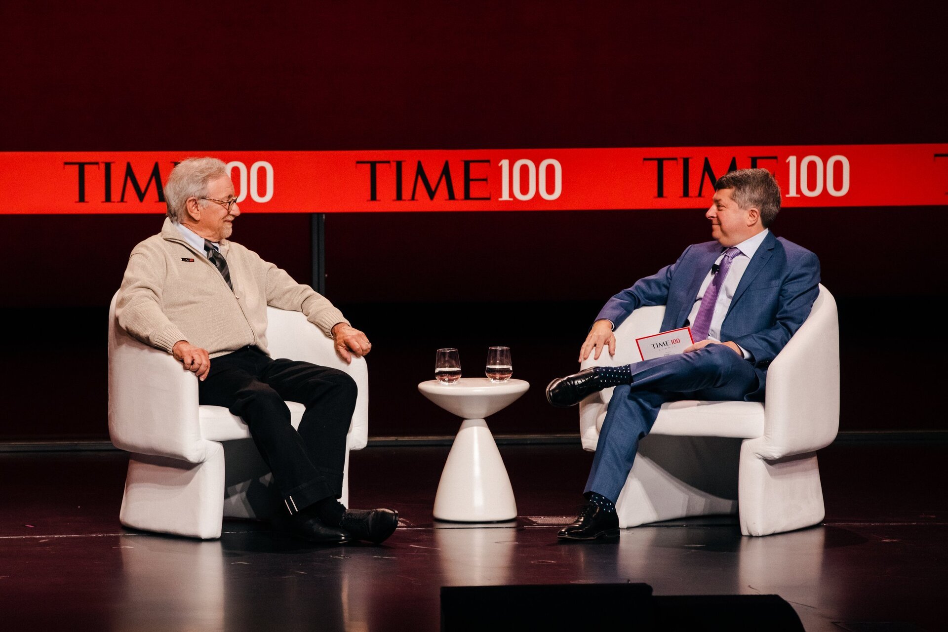 Two men in suits are sitting in chairs on a stage. A table with glasses of water sits between them. The background is red.