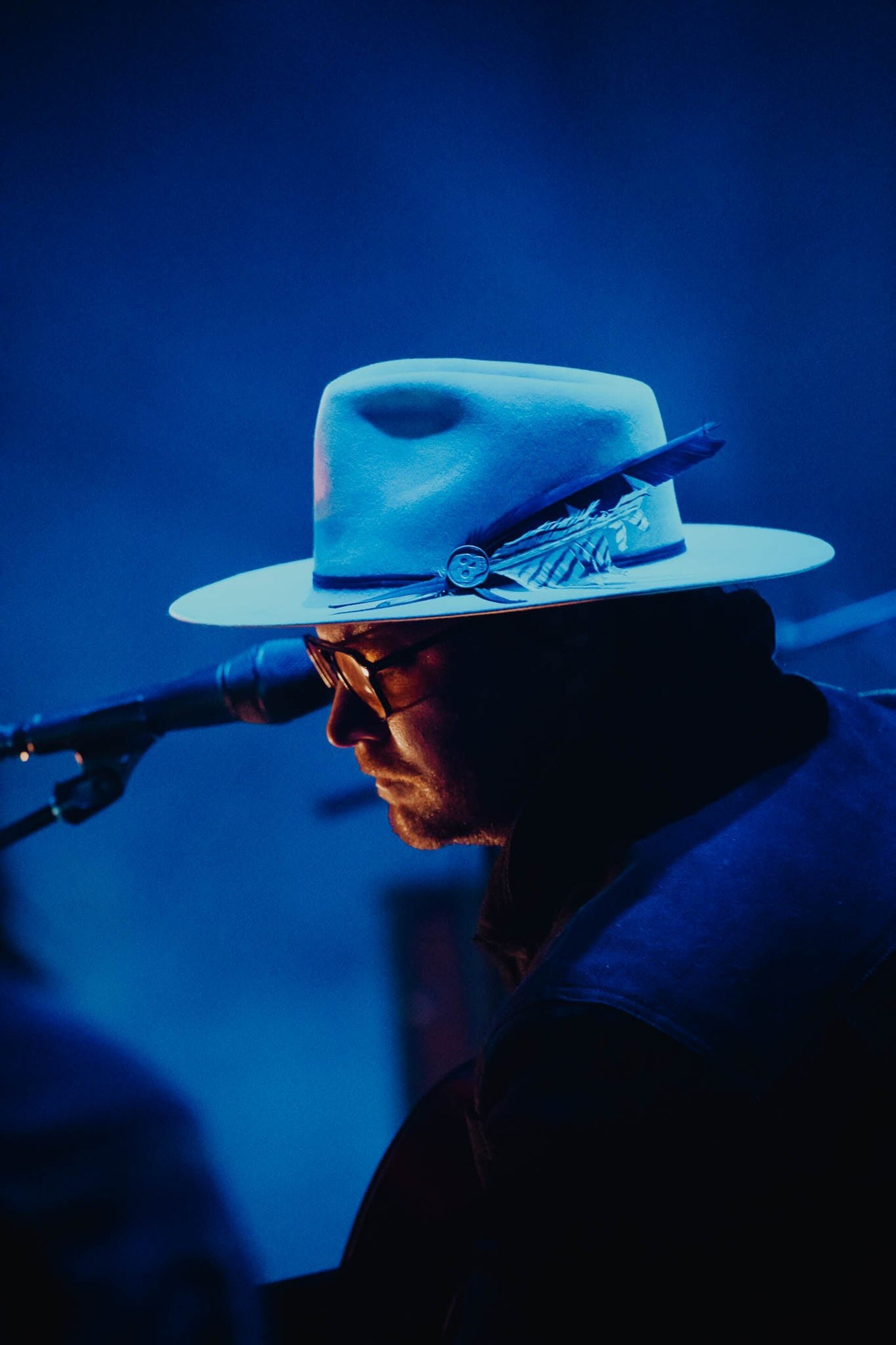 Portrait of a singer with glasses and a hat singing on a stage with a microphone in front of him bathed in blue light.