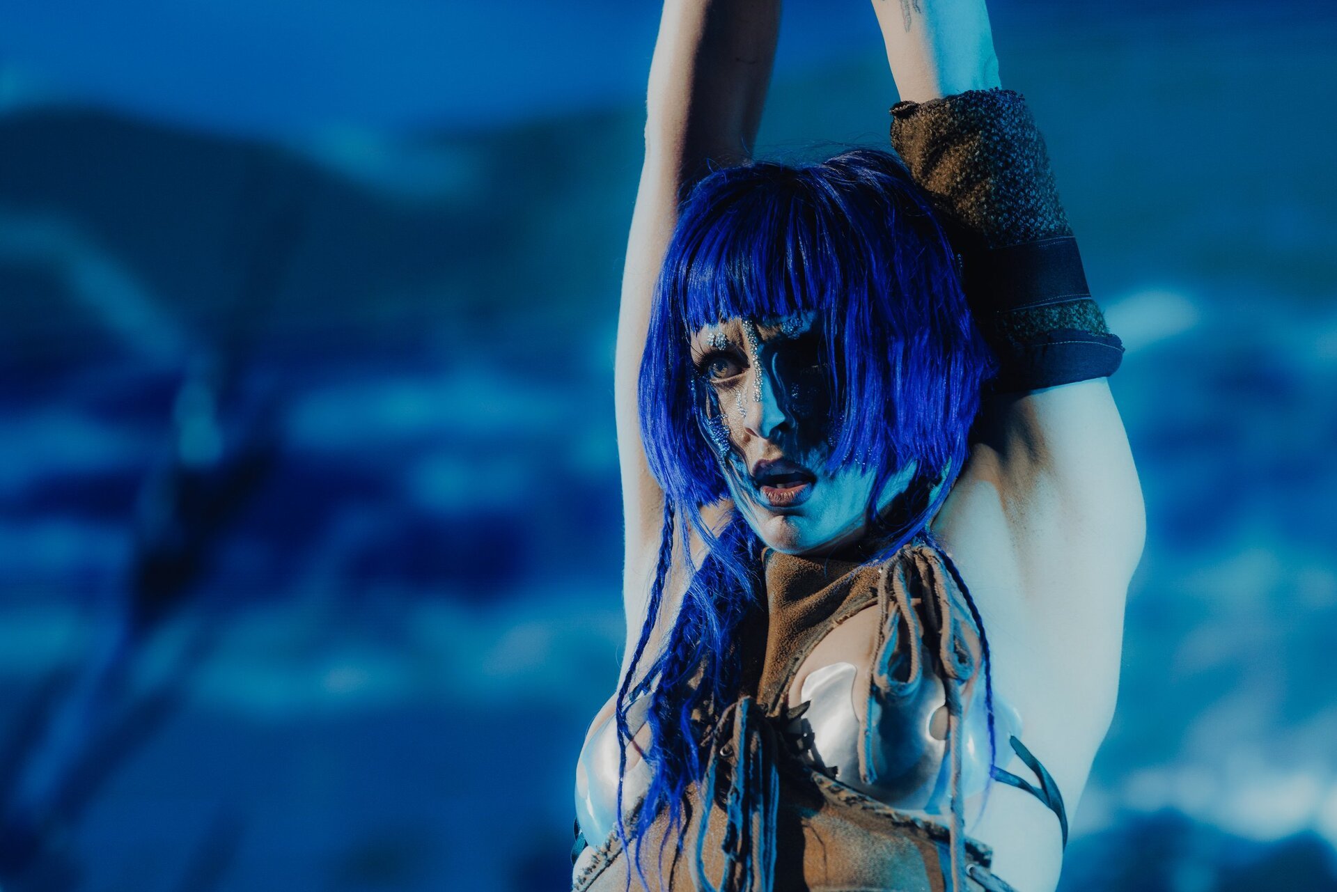 Woman with blue wig and silver body paint poses indoors against a blue cloud background, looking directly at the camera with neutral expression.
