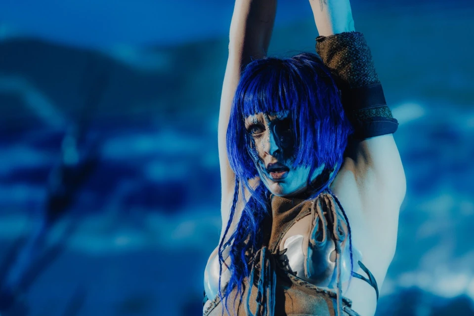 Woman with blue wig and silver body paint poses indoors against a blue cloud background, looking directly at the camera with neutral expression.