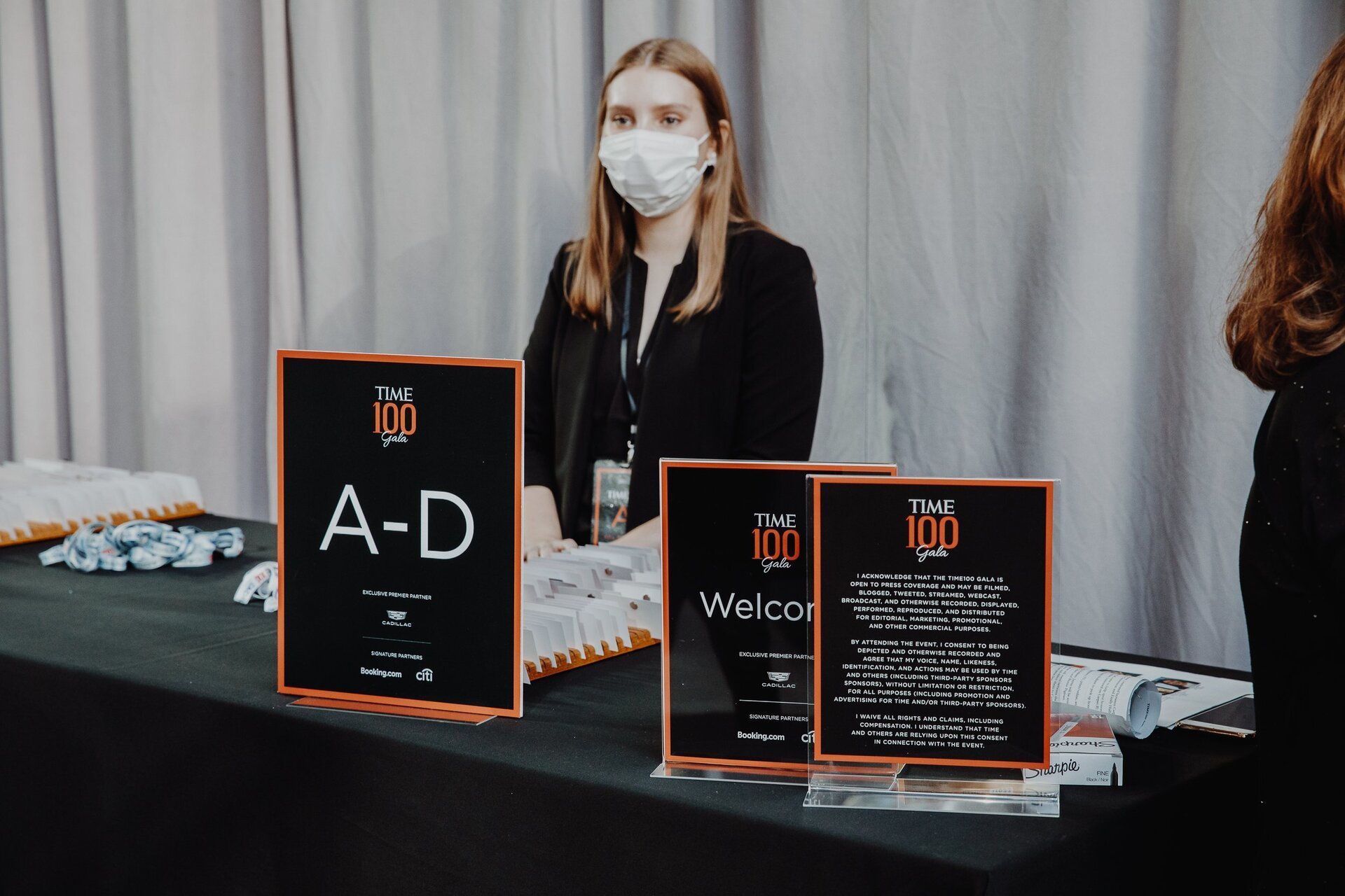 Adult female wearing a surgical mask standing behind a table with signs and materials for the Time 100 Gala event indoors.