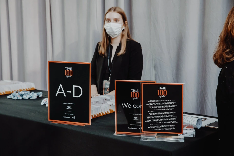 Adult female wearing a surgical mask standing behind a table with signs and materials for the Time 100 Gala event indoors.