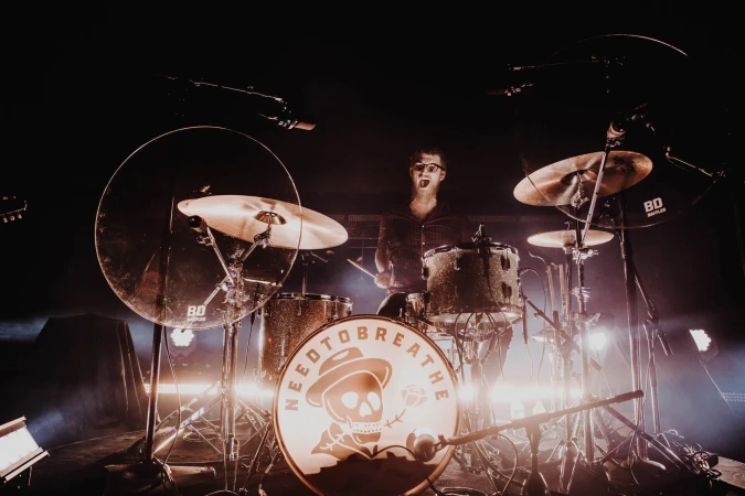 Band member plays drums in concert with the band NEEDTOBREATHE. Stage lighting highlights the drum set and singer.