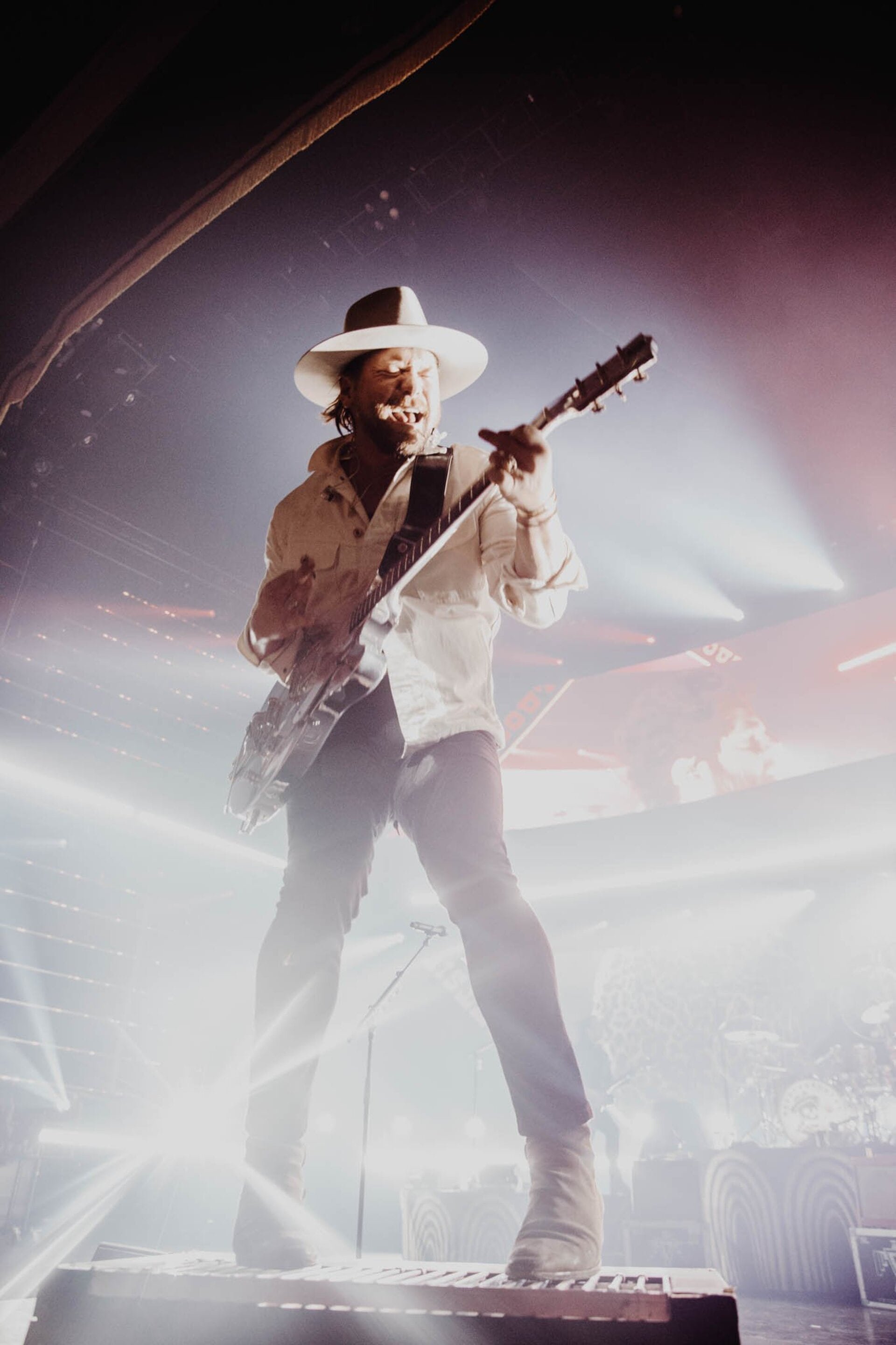 A performer in a hat and boots plays an electric guitar and sings on stage at a music concert with dramatic lighting.