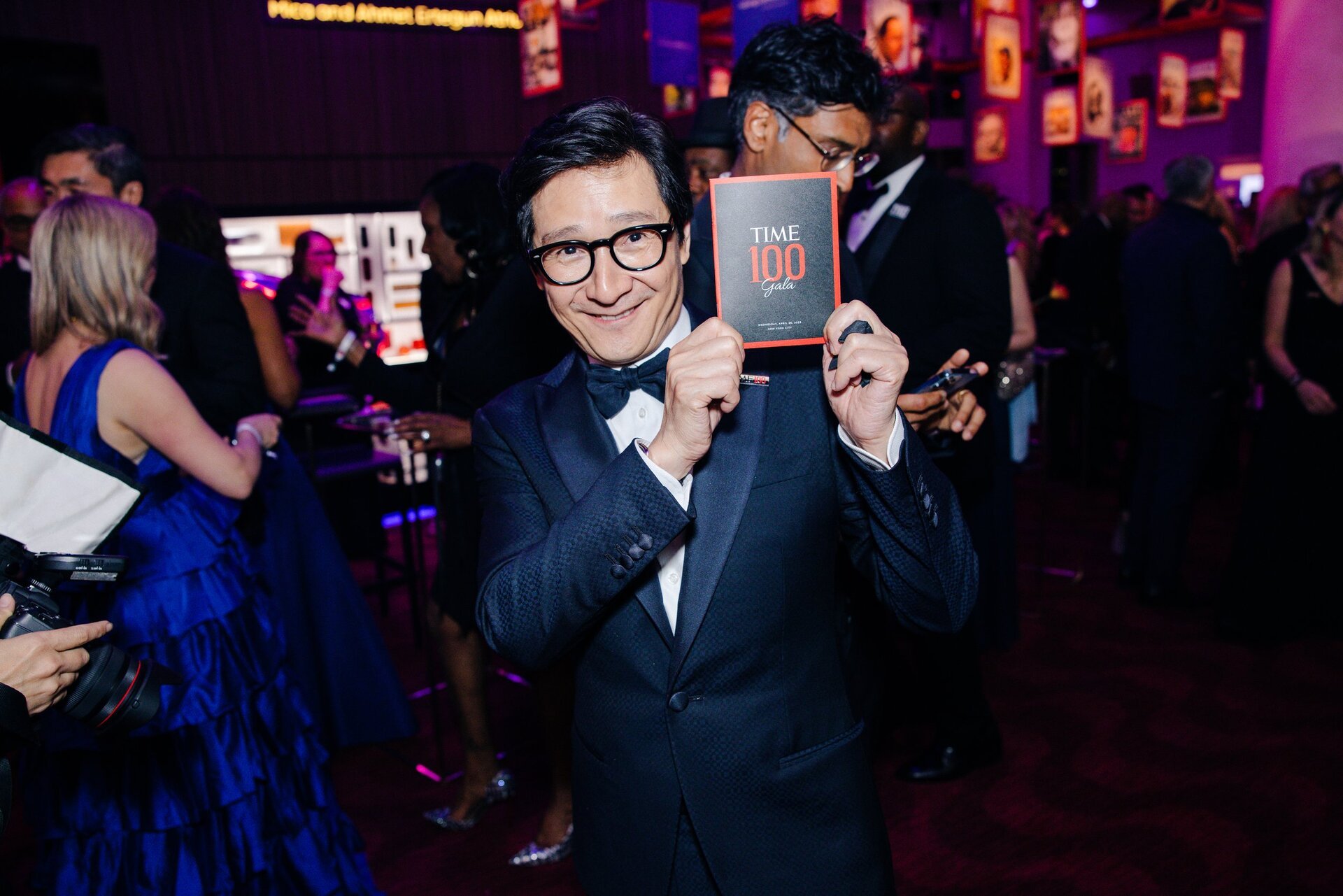 Asian man in a black tuxedo smiles while holding a Time 100 Gala card at an indoor event, captured in a medium shot.