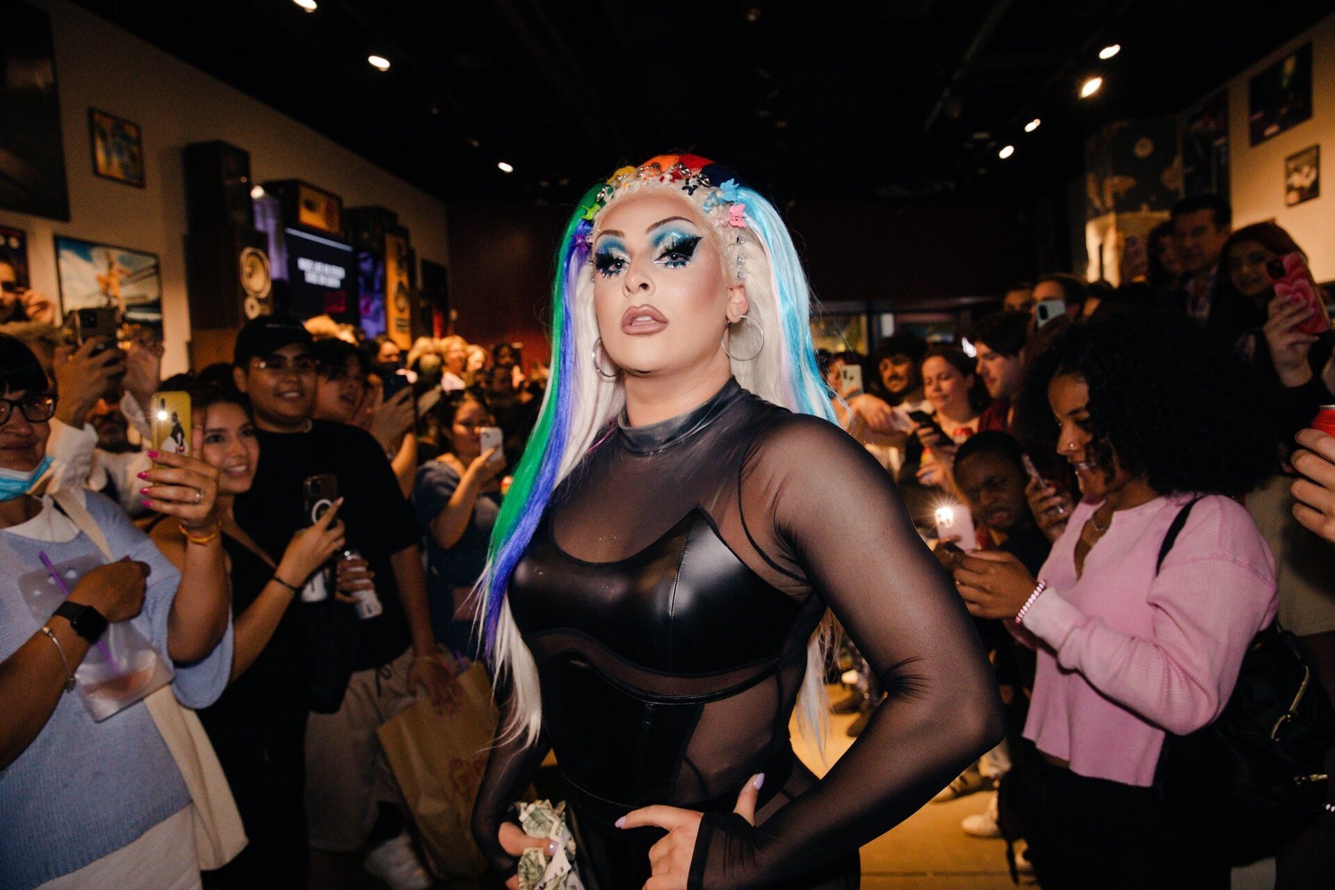 Drag queen stands out in a black bodysuit with rainbow hair, striking a pose as an audience captures her with phones in an indoor venue.