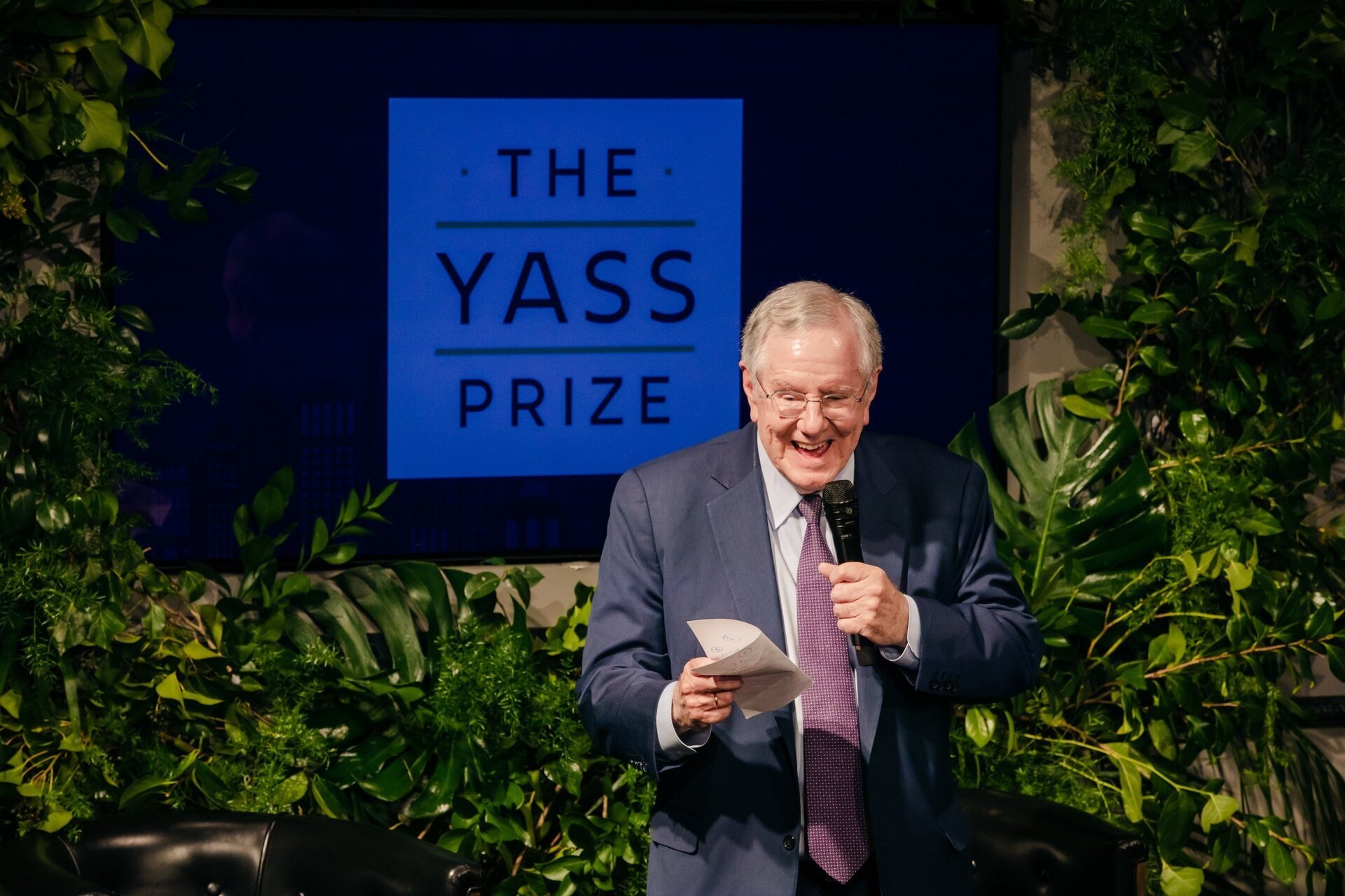 Man smiles and holds a paper and microphone during a speech in front of greenery and a screen for an event called The Yass Prize.