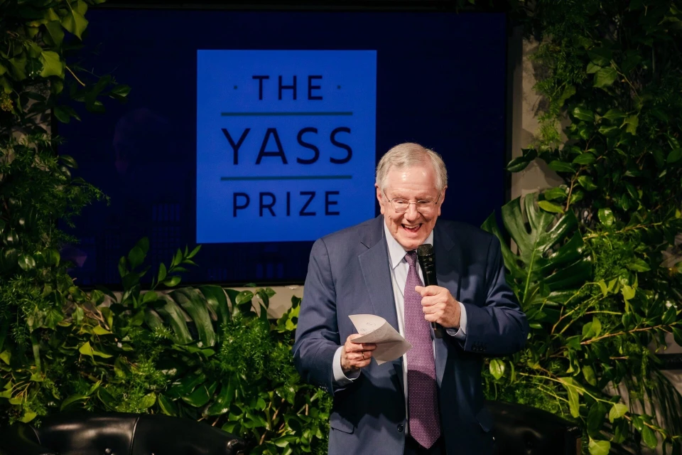 Man smiles and holds a paper and microphone during a speech in front of greenery and a screen for an event called The Yass Prize.