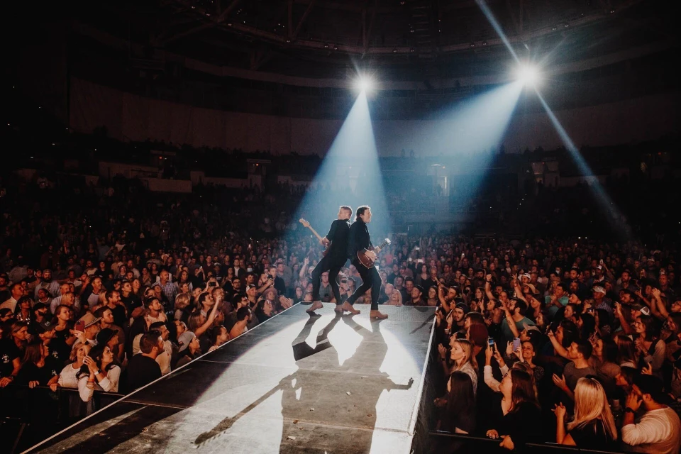 Two musicians perform on stage at a concert, bathed in bright spotlights, with a large crowd of fans in the background.