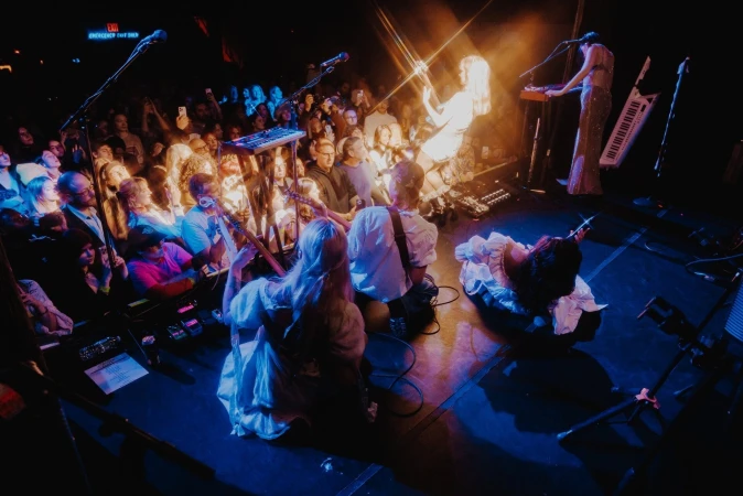Live music performance on stage with a band, musicians, instruments, and a large audience in a darkened concert venue with vibrant lighting.