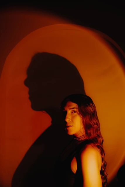 Portrait of a young adult with long wavy hair, posing in a photo studio under orange lighting with a large head shadow.