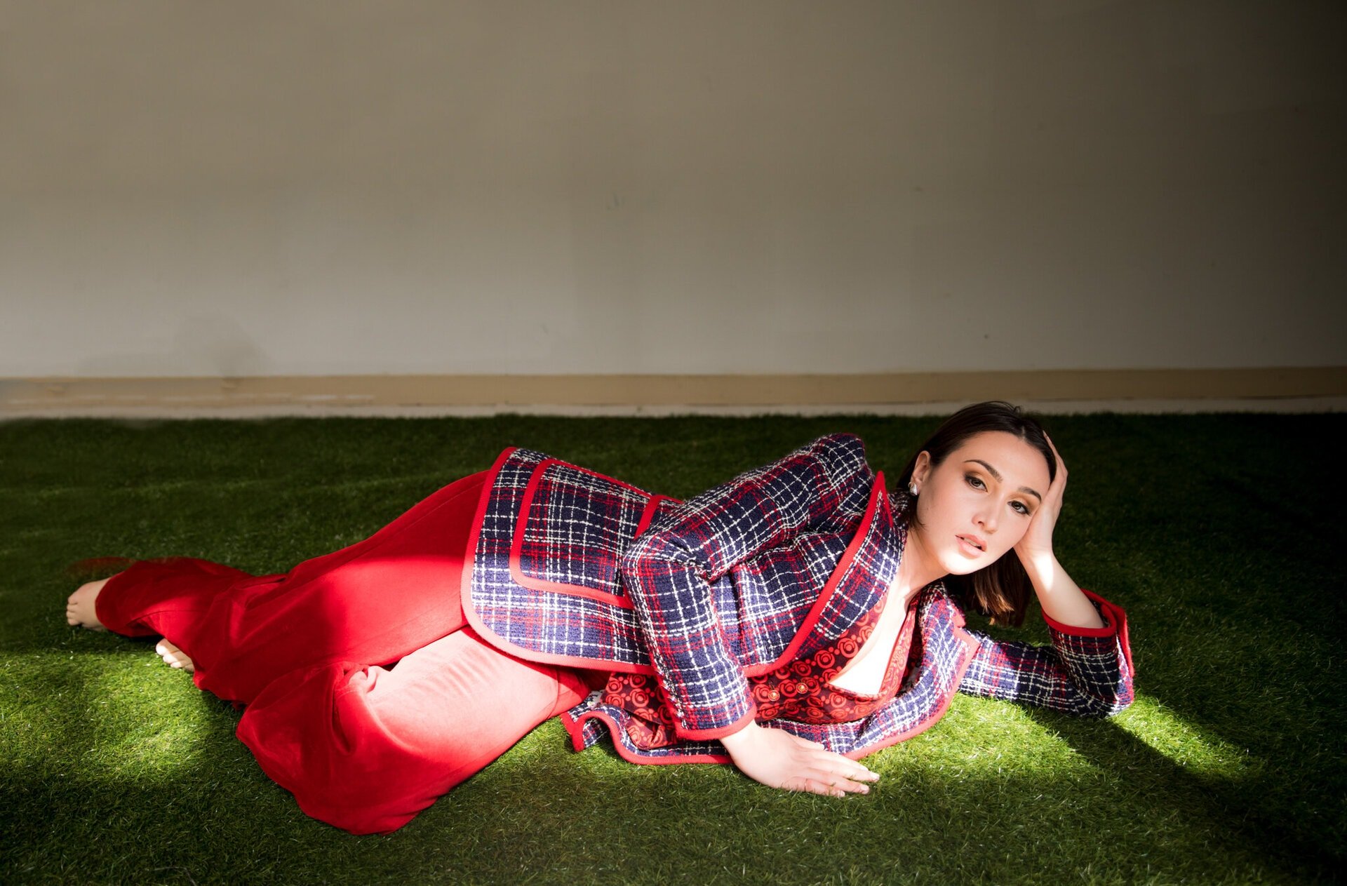 Woman in a plaid blazer and red pants lies on artificial grass, gazing at the camera. Sunlight illuminates her face and outfit.