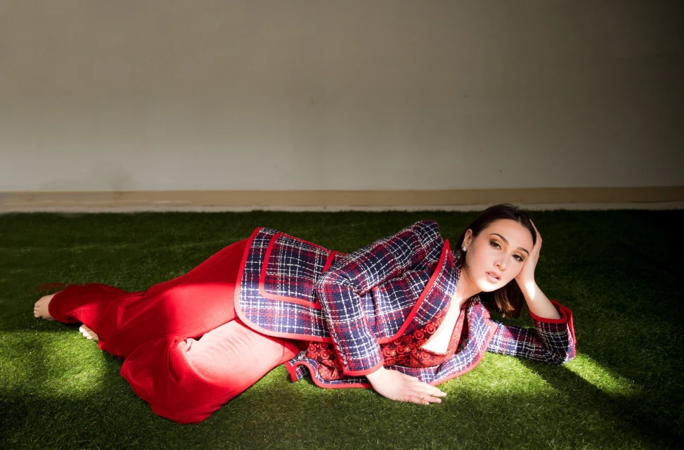 Woman in a plaid blazer and red pants lies on artificial grass, gazing at the camera. Sunlight illuminates her face and outfit.