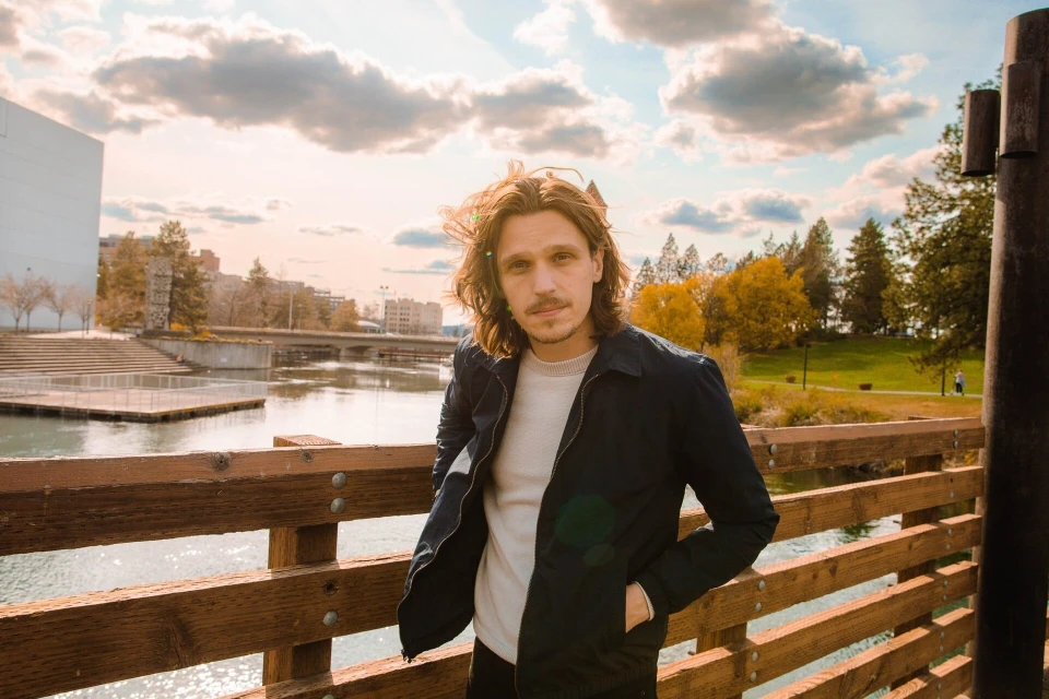 An adult male with long hair poses on a wooden bridge with a river, park, and buildings in the background under a bright, cloudy sky.
