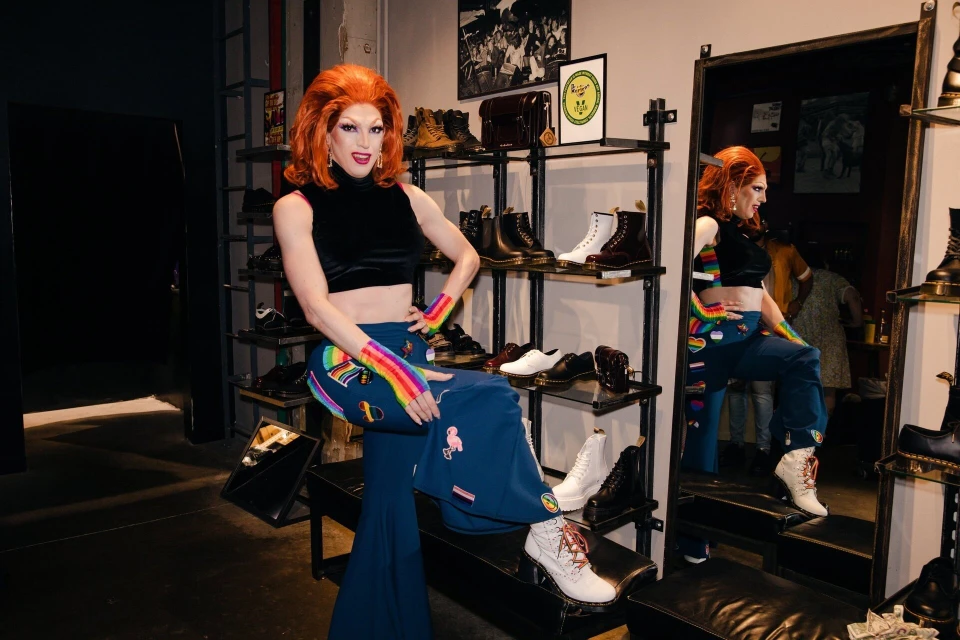 Drag queen with orange hair poses in a retail setting, showcasing a black top and rainbow-themed jeans and arm warmers near shoe shelves.