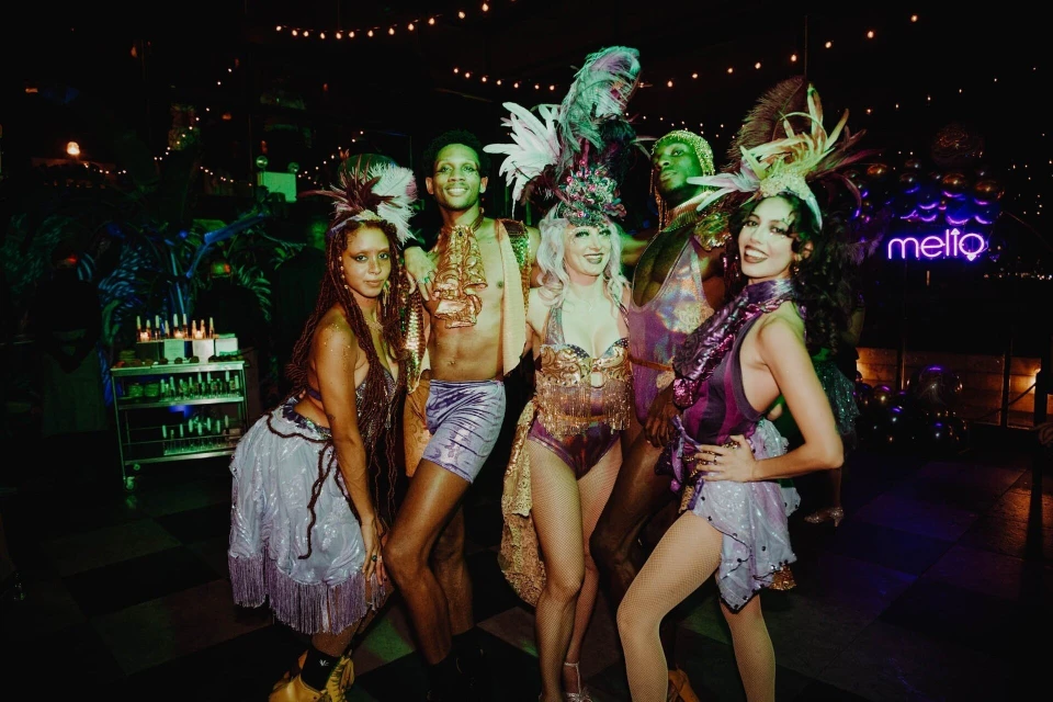 Group of performers in theatrical costumes and feather headdresses pose on a stage with disco lighting in a dimly lit interior venue.
