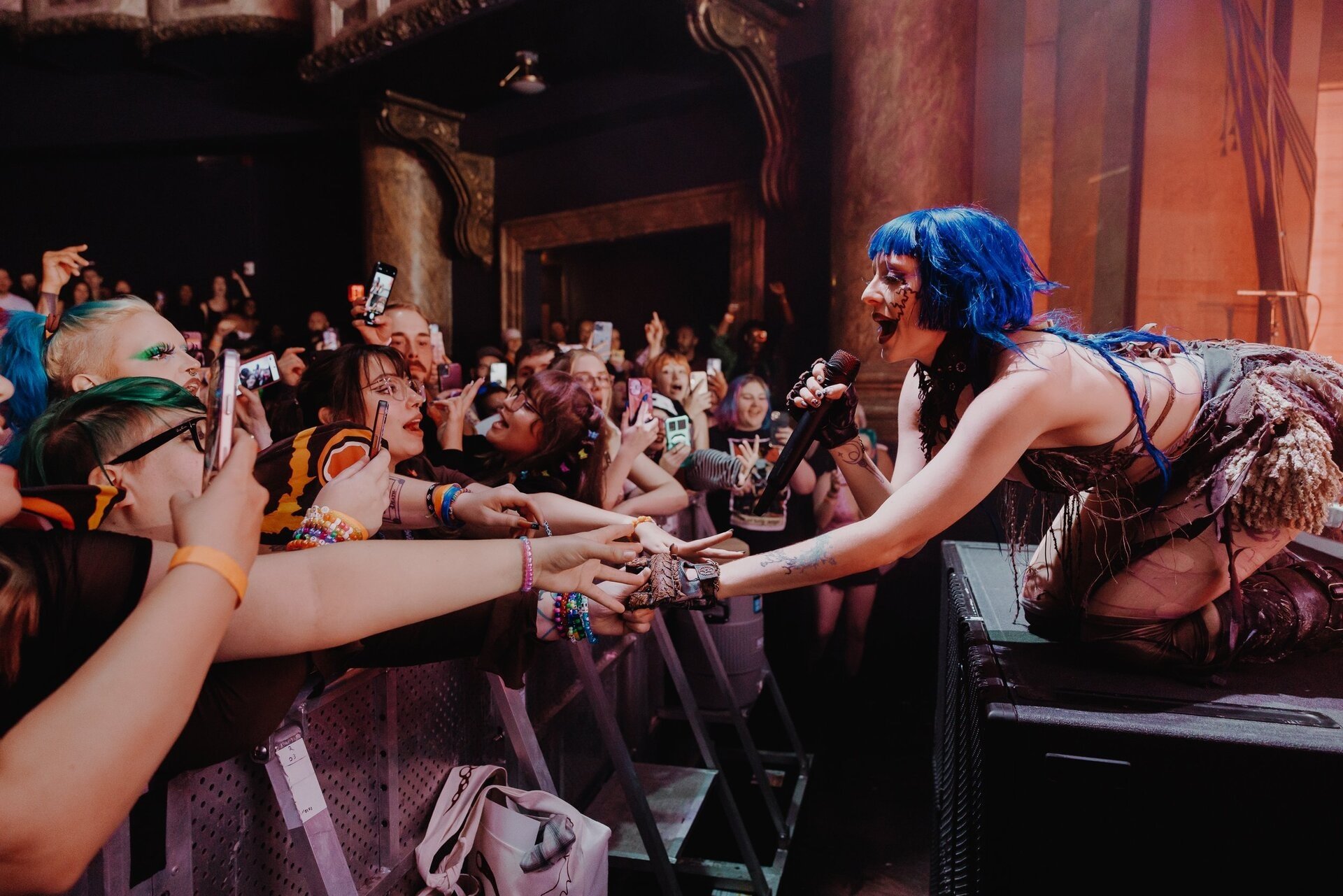 A female musician with blue hair kneels on a stage performing to a large crowd holding up phones in a concert venue with warm lighting.