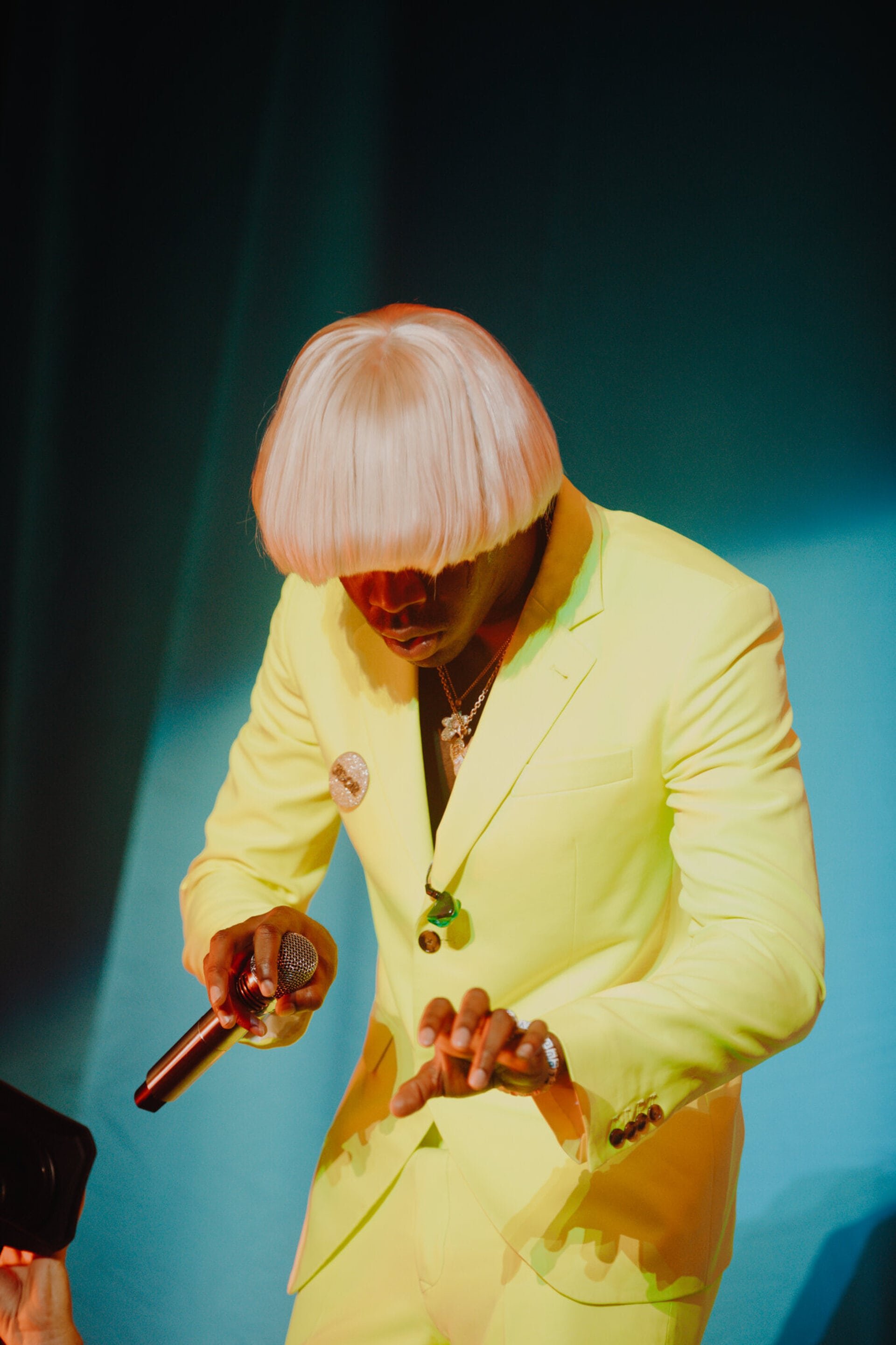 Concert photo of a singer with a blonde wig in a yellow suit holding a microphone, performing on stage with a blue background.