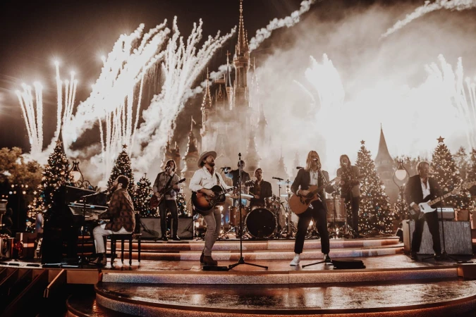 Band performs live on stage with Christmas trees and fireworks, backed by fairytale castle at nighttime concert event, shot at eye-level.