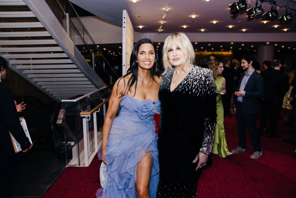 Two women pose on a red carpet in front of a staircase and crowd. One wears a light blue dress, and the other wears a black gown.