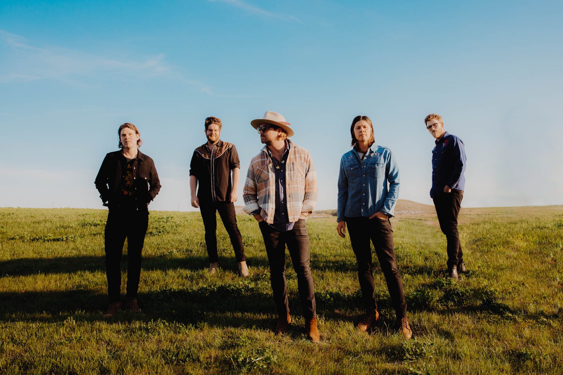 Five adult men stand in an outdoor grassy field under a blue sky, wearing casual clothing, boots, and a hat, posing for a photo.
