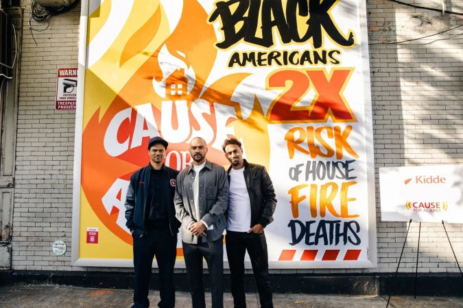 Three adult males standing in front of a public service announcement billboard with black, orange, and white text and graphic elements.