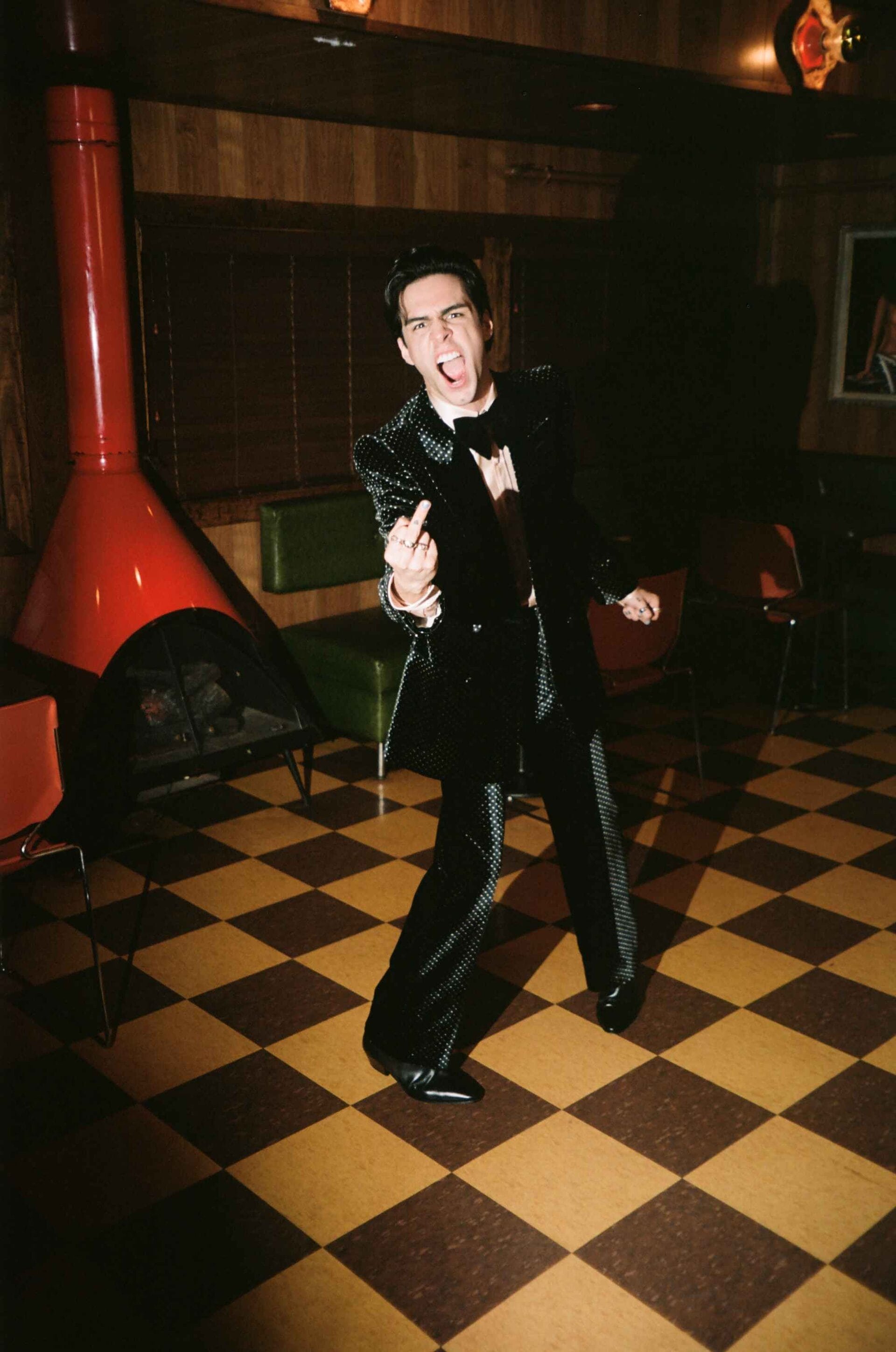 A man in a sequined suit stands on a checkerboard floor with a vintage fireplace and wood paneling, sticking out his tongue and gesturing.